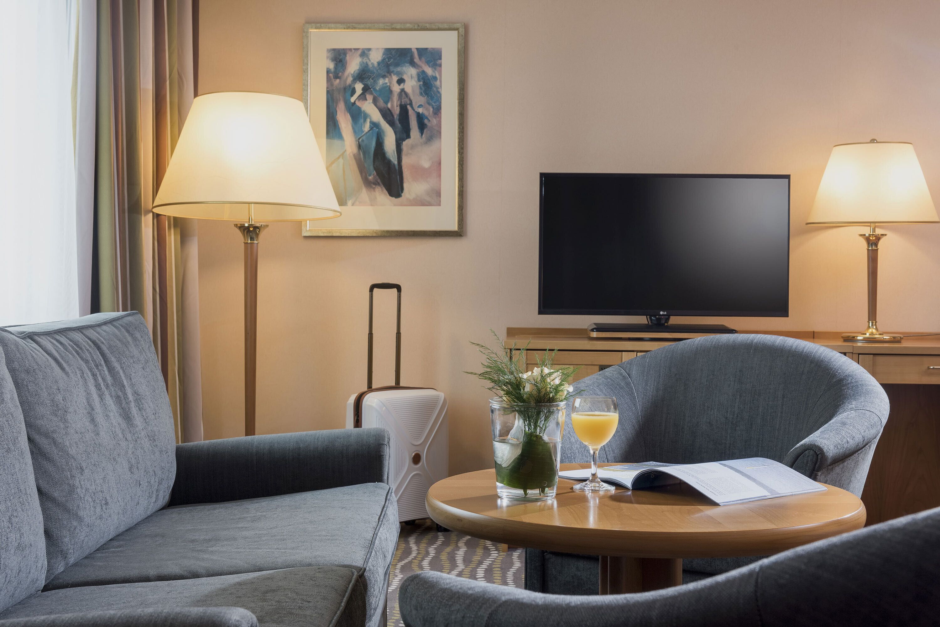 Habitación de hotel con sofá, sillón y una mesita con un vaso de zumo de naranja y un libro. Hay un televisor de pantalla plana sobre un mueble bajo y una lámpara a cada lado del televisor.