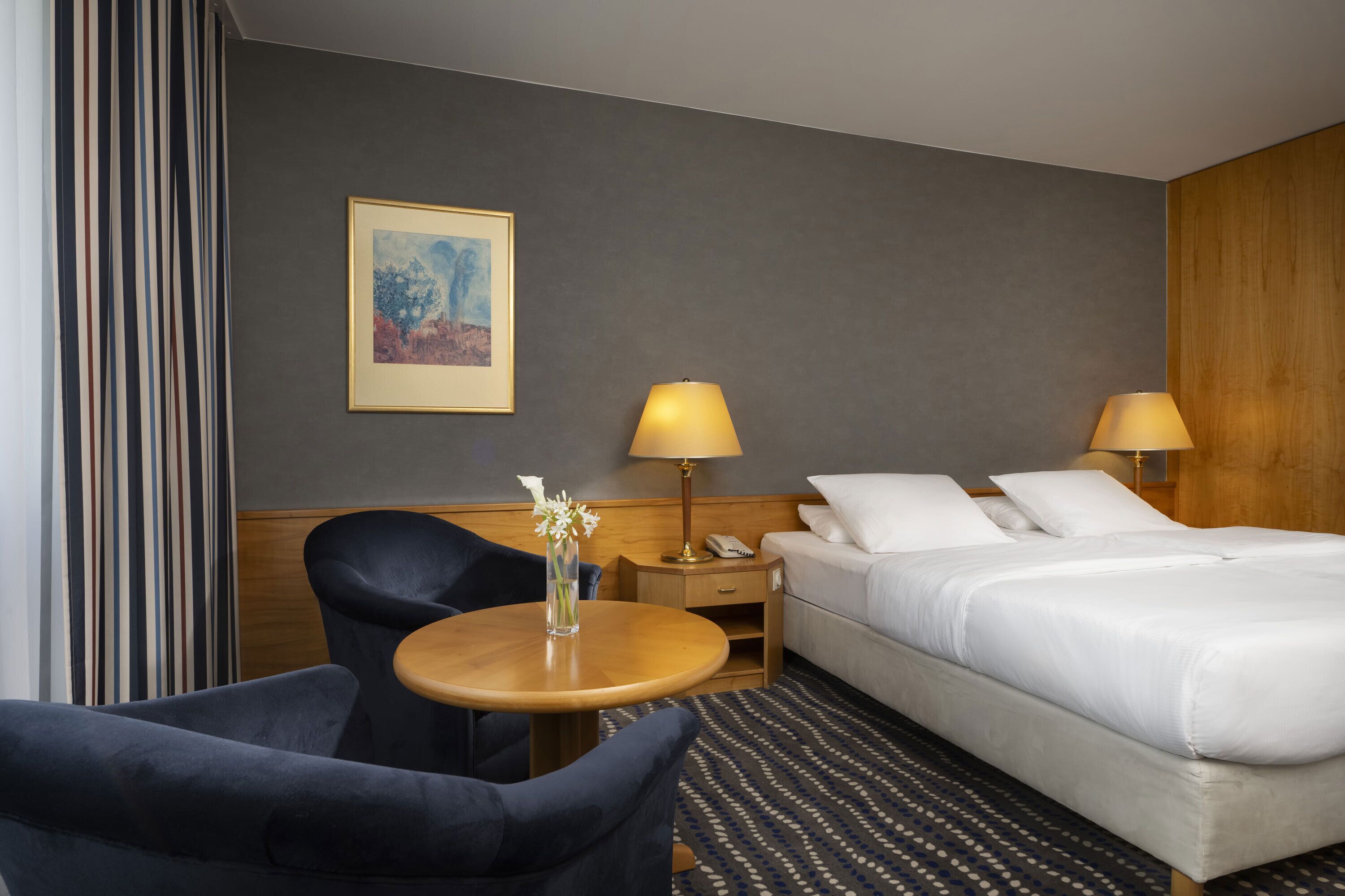 Une chambre d’hôtel avec un lit, deux lampes de chevet, une petite table avec un vase, une chaise, une table de chevet avec un téléphone et un tableau au mur.