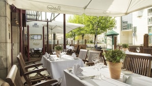 An outdoor dining area with tables set for two, covered by white umbrellas, and wooden chairs. - Maritim Hotel Magdeburg (Magdeburg)