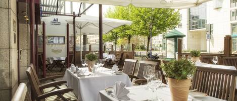 An outdoor dining area with tables set for two, covered by white umbrellas, and wooden chairs.