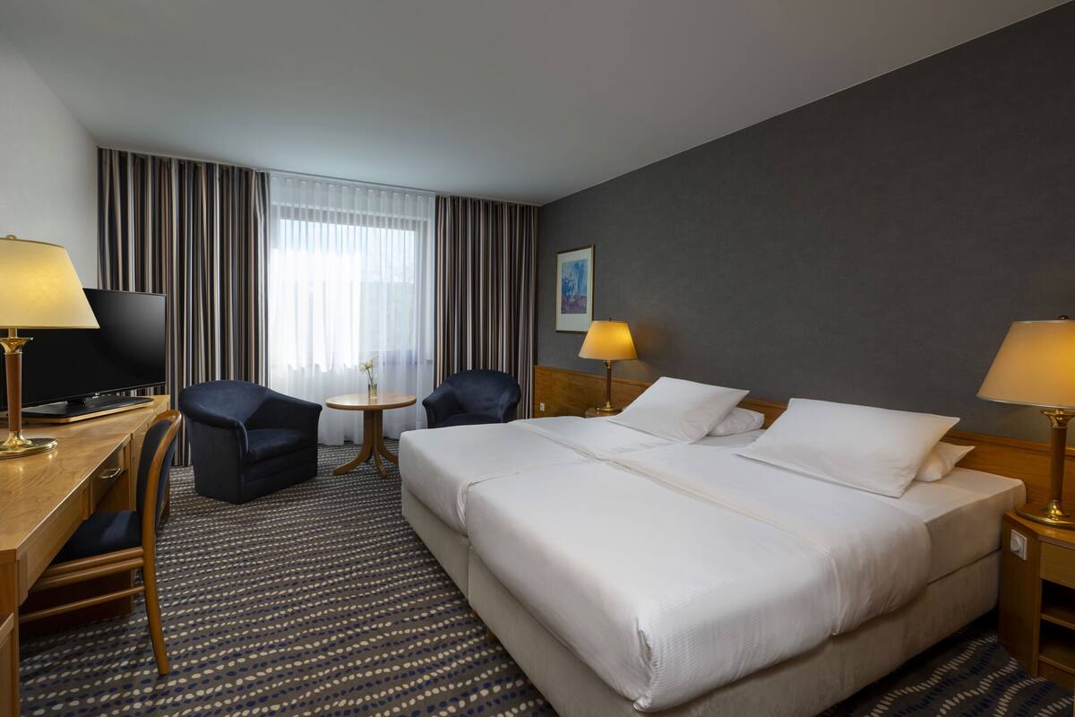 A hotel room with a large bed, two chairs, a desk with a TV, and a small table with a lamp.