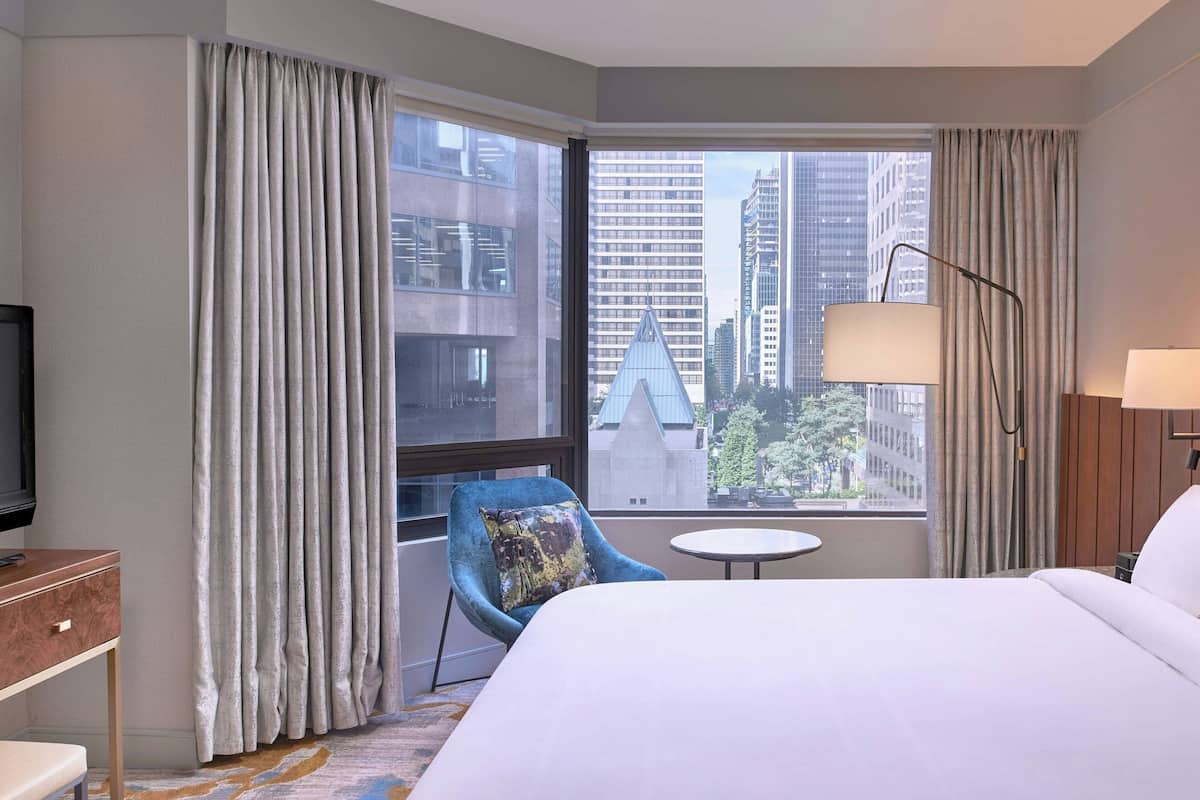 Una habitación de hotel con una cama grande, un sillón azul, una mesita pequeña, un televisor y vistas a edificios de la ciudad.