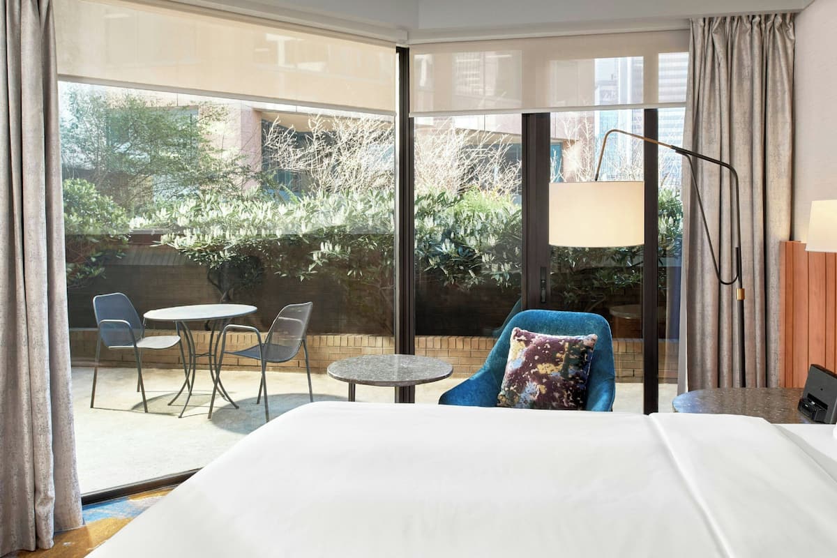 Habitación de hotel con una cama grande, un sillón azul, una mesita redonda y vistas a un jardín a través de amplios ventanales.