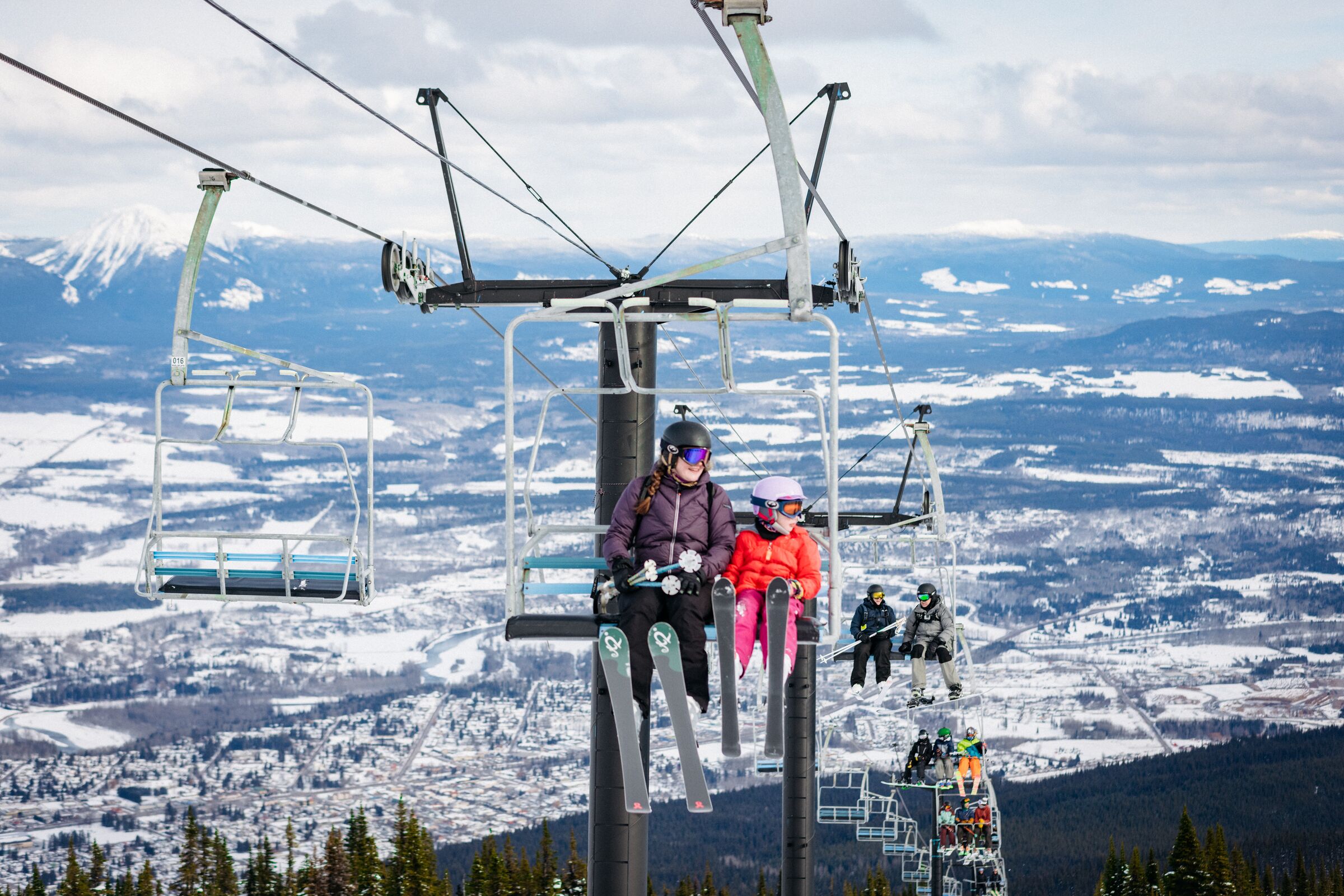 A ski lift with skiers seated on it, overlooking a snowy landscape with mountains and a town below.