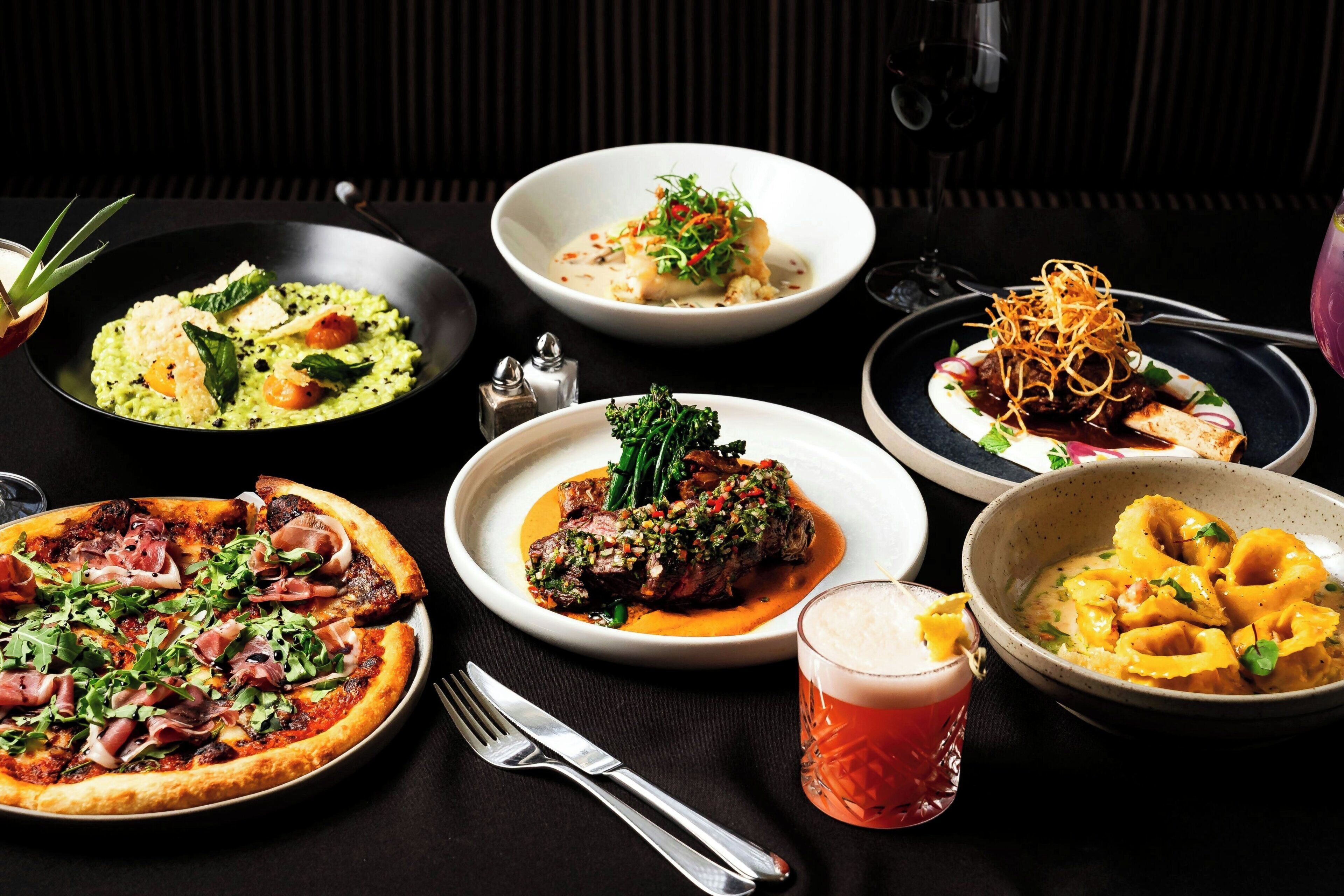 A variety of dishes on a table, including a pizza, a bowl of soup, and a plate with meat and vegetables.