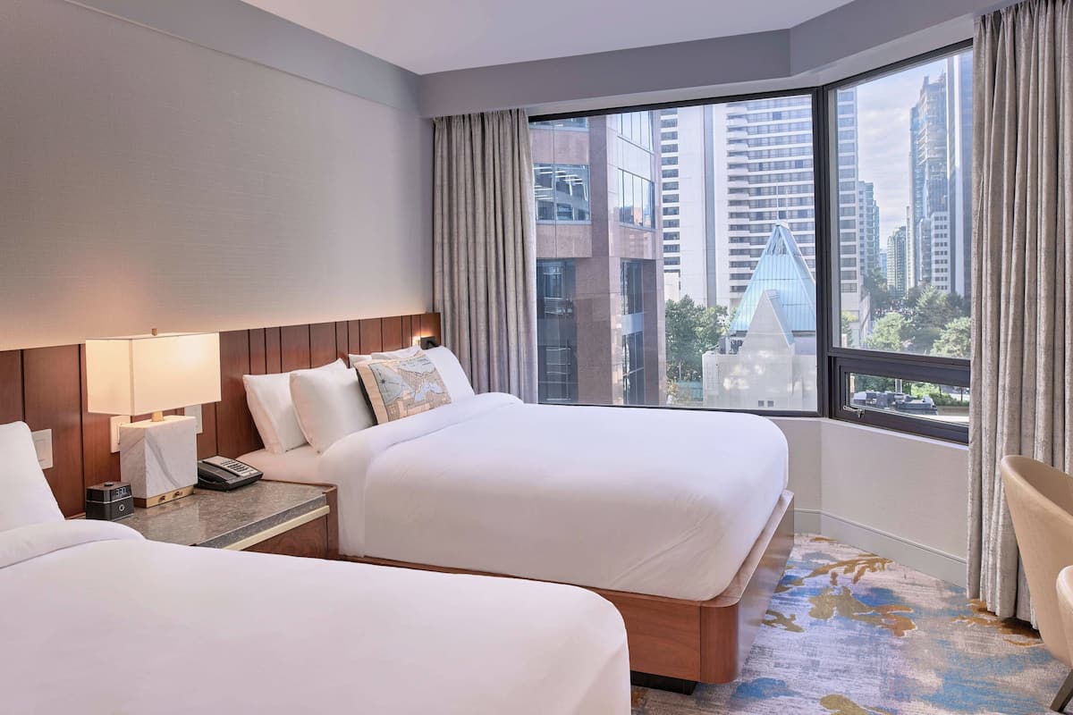 Habitación de hotel con dos camas, una mesita de noche, una lámpara y vistas de la ciudad a través de amplias ventanas.