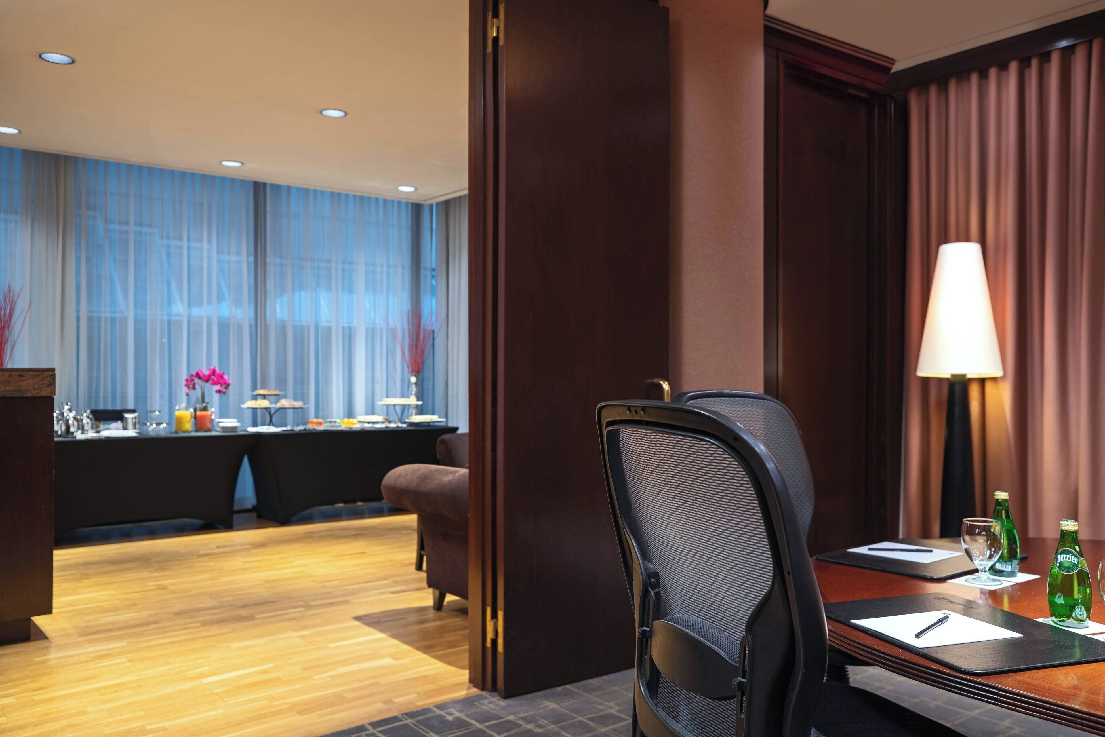Habitación de hotel con suelo de madera, una mesa de comedor con sillas, una lámpara y vistas a un comedor con una mesa dispuesta para comer.
