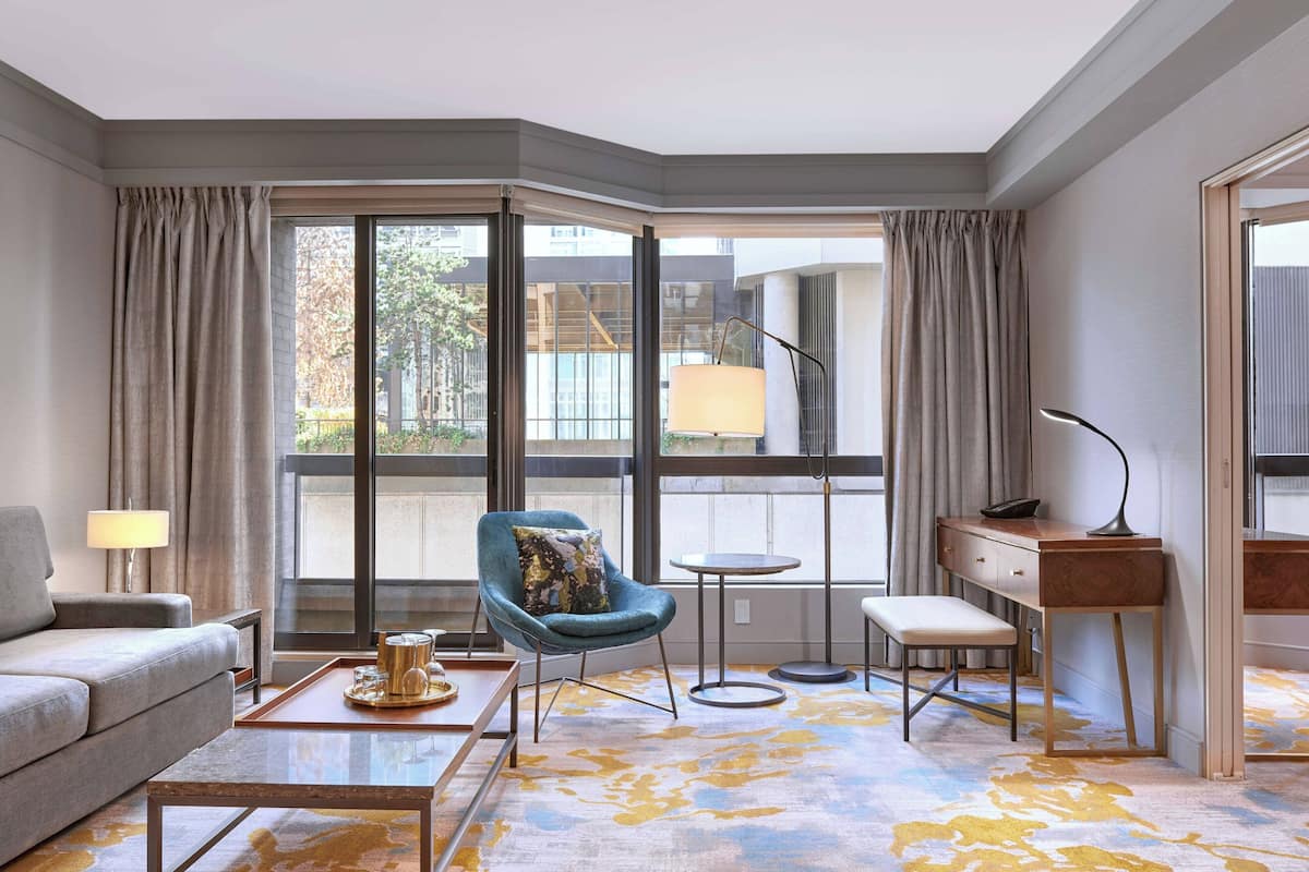 Una habitación de hotel moderna con un sofá, una mesa de centro, un escritorio y una silla. Cuenta con un ventanal con cortinas, una lámpara y vistas al exterior.