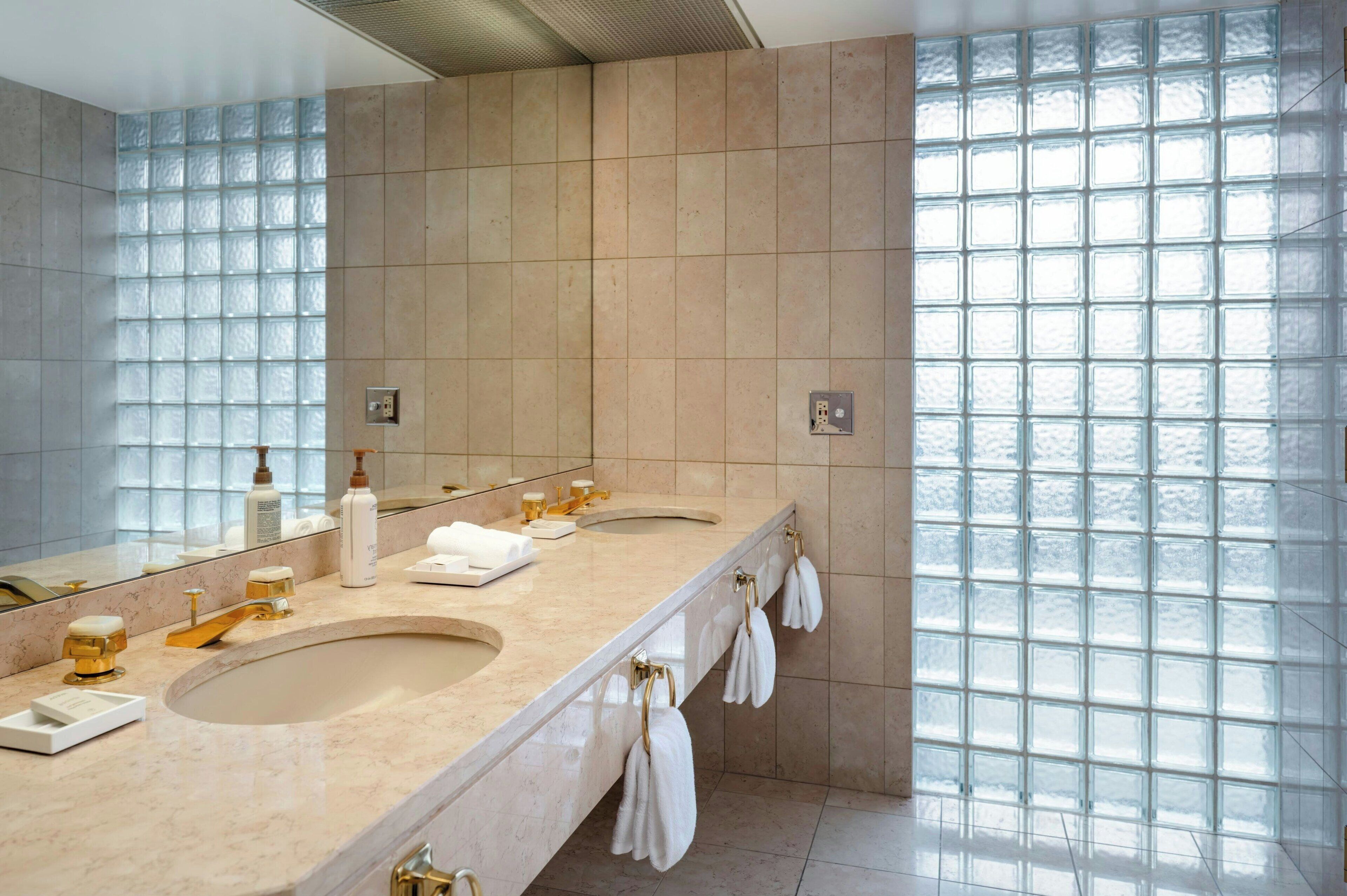 A modern bathroom with two sinks, a large mirror, and a view through a glass block wall.