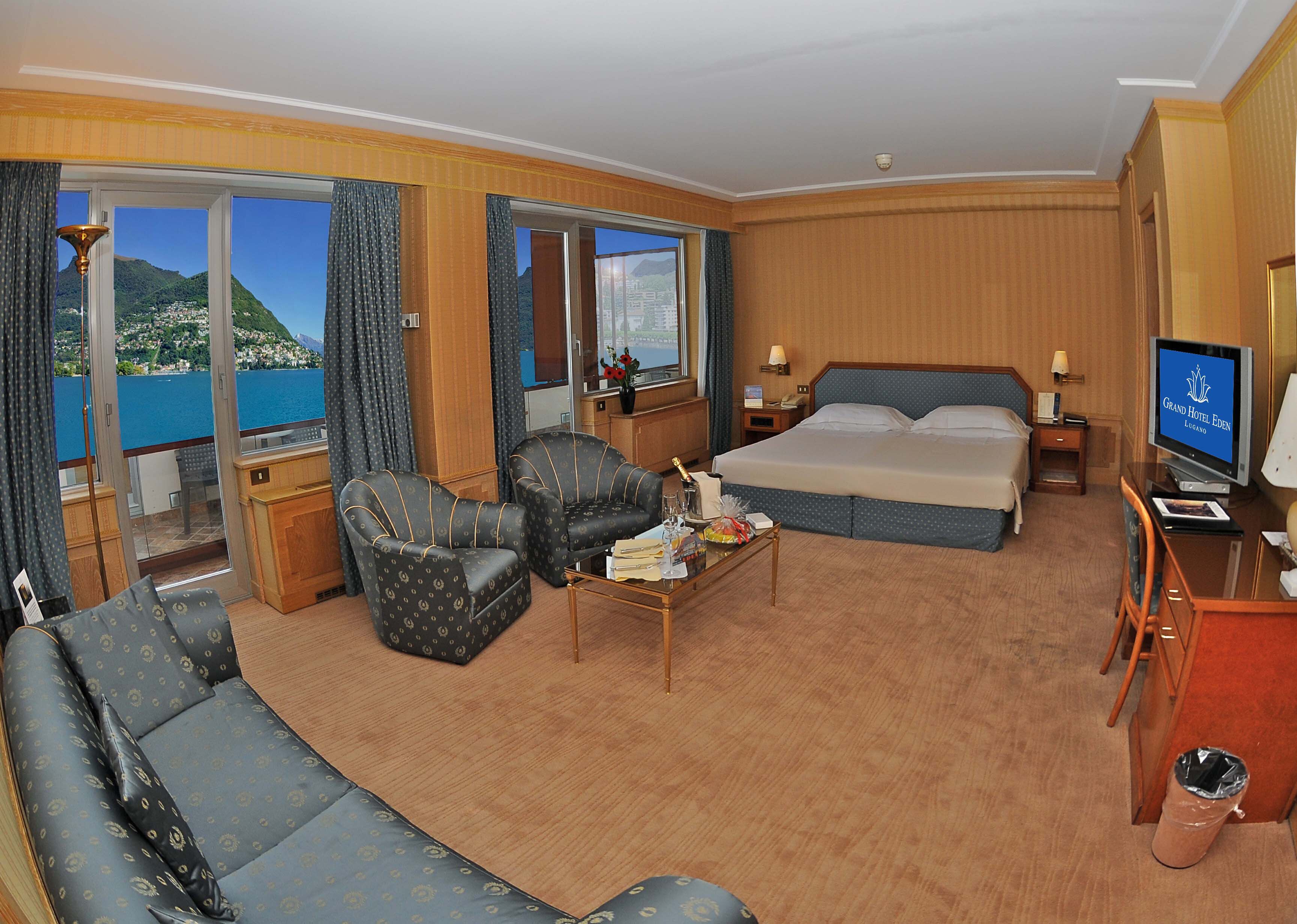 Une chambre d’hôtel comprenant un lit, deux fauteuils, un canapé, une petite table avec une bouteille et des verres, une télévision, et une vue sur un plan d’eau et des montagnes.