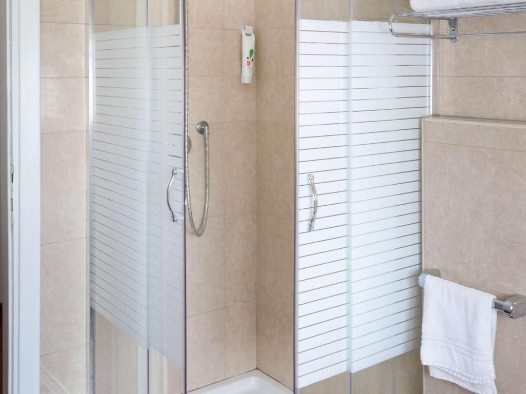 Area doccia vetrata con soffione a mano, porta asciugamani con asciugamano bianco e dispenser per sapone a parete.