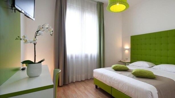 A hotel room with a green upholstered headboard, a bed with white bedding and green pillows, a bedside table with a lamp, and a potted orchid on a white table. - Hotel Milano Palmanova (Milan)