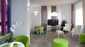 A modern living room with a flat-screen TV, green and white chairs, a small table with a plant, and a large window. - Hotel Milano Palmanova (Milan)