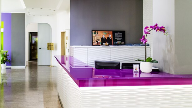 A reception area with a purple counter, a digital display, and a potted orchid. - Hotel Milano Palmanova (Milan)