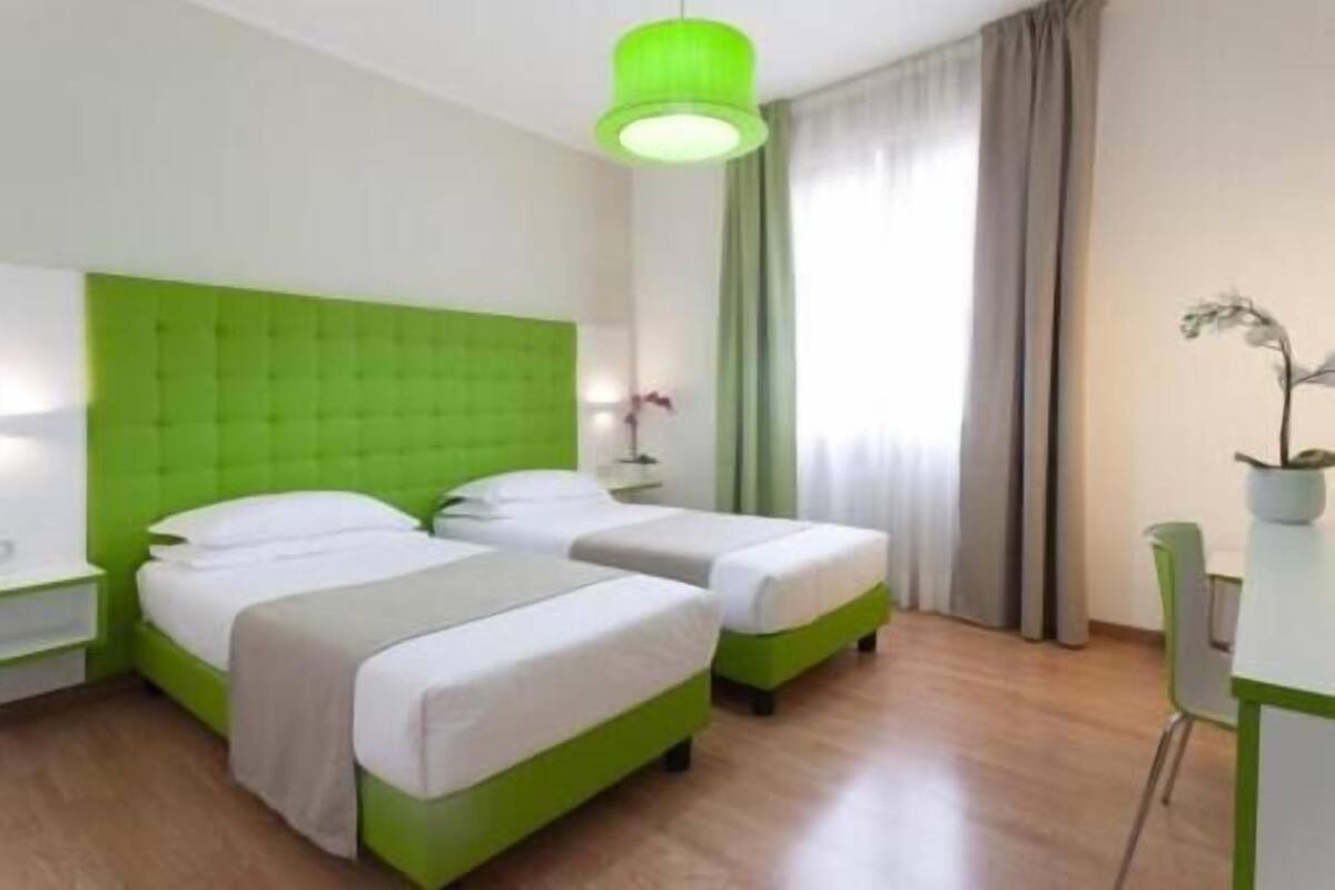 Hotellihuone, jossa on kaksi sänkyä, vihreä pääty, yöpöytä lampun kanssa, ruukkukasvi ja ikkuna, jossa on verhot.
