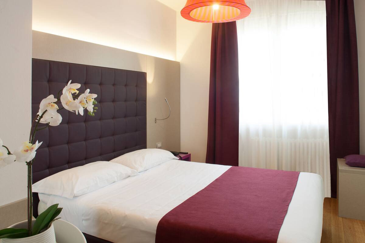 Moderni hotellihuone, jossa on suuri sänky, yöpöytä, yövalaisin, ruukkukasvi ja ikkuna, jossa on verhot.