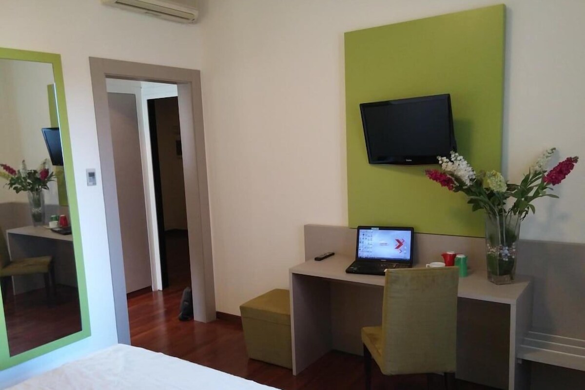 Ein Hotelzimmer mit Bett, Schreibtisch, Laptop, Fernseher, Stuhl und Blumen in einer Vase.