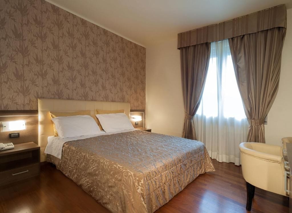 Une chambre d’hôtel avec un grand lit, une table de chevet avec une lampe, une chaise et une fenêtre avec des rideaux.