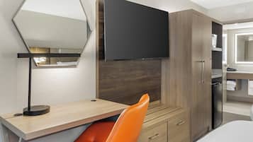 A hotel room with a bed, a desk with a chair, a flat-screen TV mounted on the wall, a wardrobe, and a bathroom with a mirror.