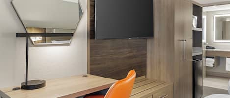 A hotel room with a bed, a desk with a chair, a flat-screen TV mounted on the wall, a wardrobe, and a bathroom with a mirror.
