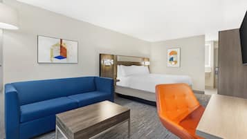 A hotel room with a bed, a blue sofa, an orange chair, a wooden coffee table, and artwork on the walls.