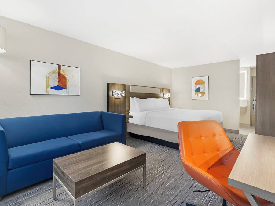 A hotel room with a bed, a blue sofa, an orange chair, a wooden coffee table, and artwork on the walls.