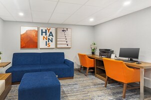 Habitación de hotel con sofá azul, una silla naranja, escritorio con computadora y obras de arte en la pared.
