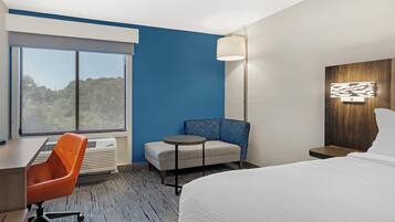 Habitación de hotel con cama, escritorio, una silla naranja, una mesita redonda pequeña, un sofá azul, una lámpara de pared y una ventana con vista a árboles.