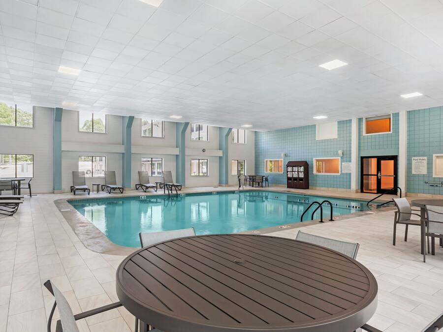 An indoor swimming pool with lounge chairs and tables, large windows, and a tiled wall.