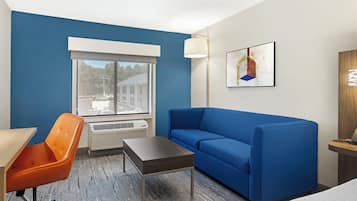 Habitación de hotel con sofá azul, sillón naranja, una mesita y ventana con vistas a edificios, además de un cuadro en la pared.