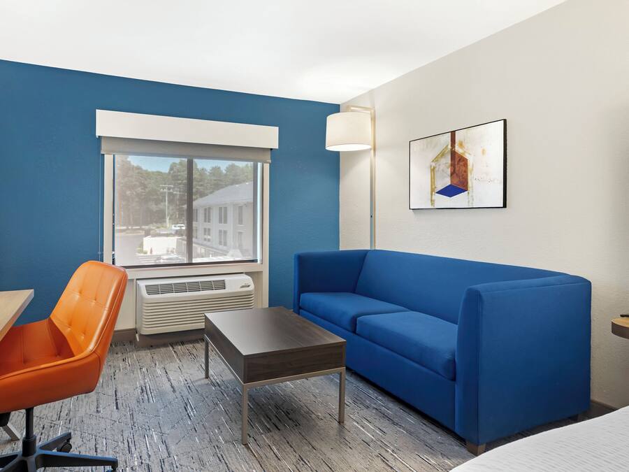Ein Hotelzimmer mit einem blauen Sofa, einem orangefarbenen Sessel, einem kleinen Tisch, einem Fenster mit Blick auf Gebäude und einem Bild an der Wand.