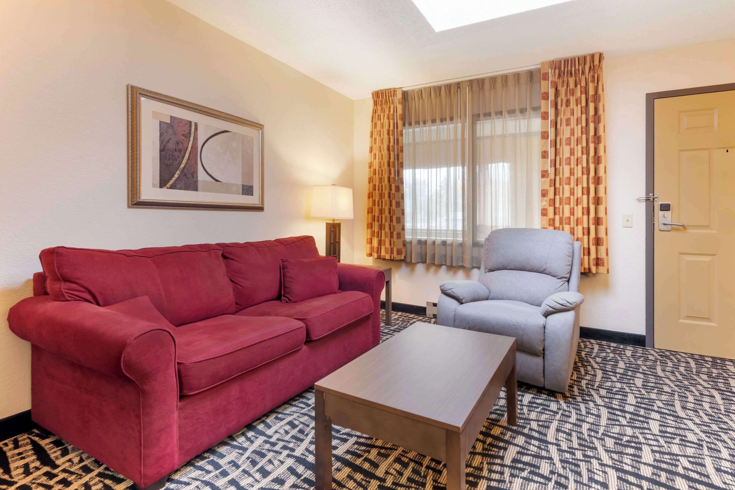 A hotel room with a red sofa, a grey armchair, a wooden coffee table, a patterned carpet, a window with curtains, a wall-mounted lamp, and a framed picture.
