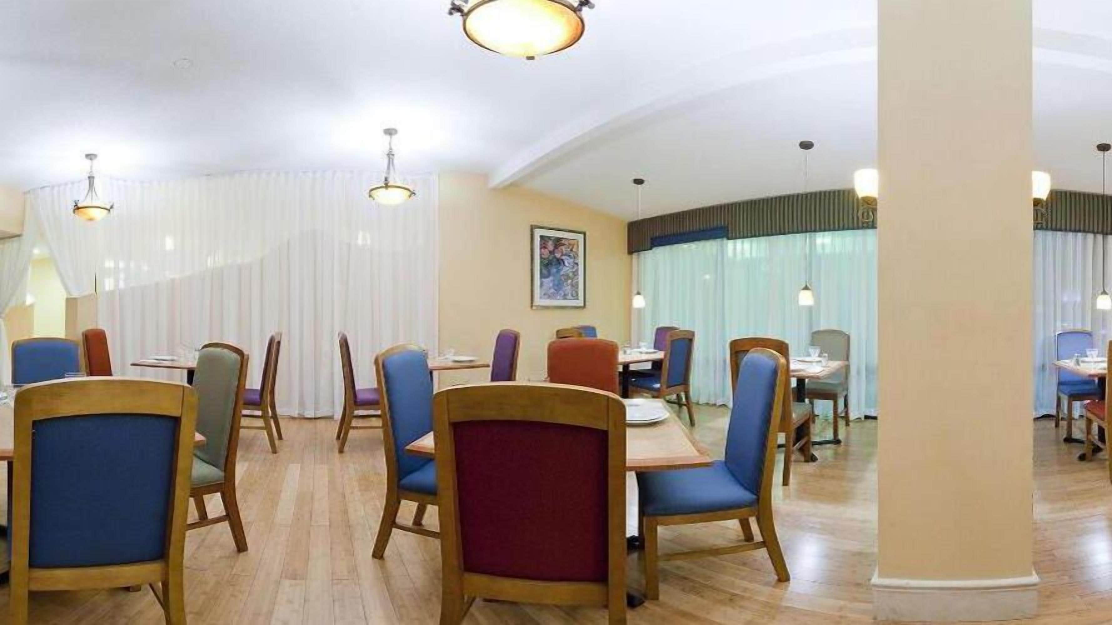 En hotellrestaurang med trästolar och bord, vita gardiner och hängande lampor.