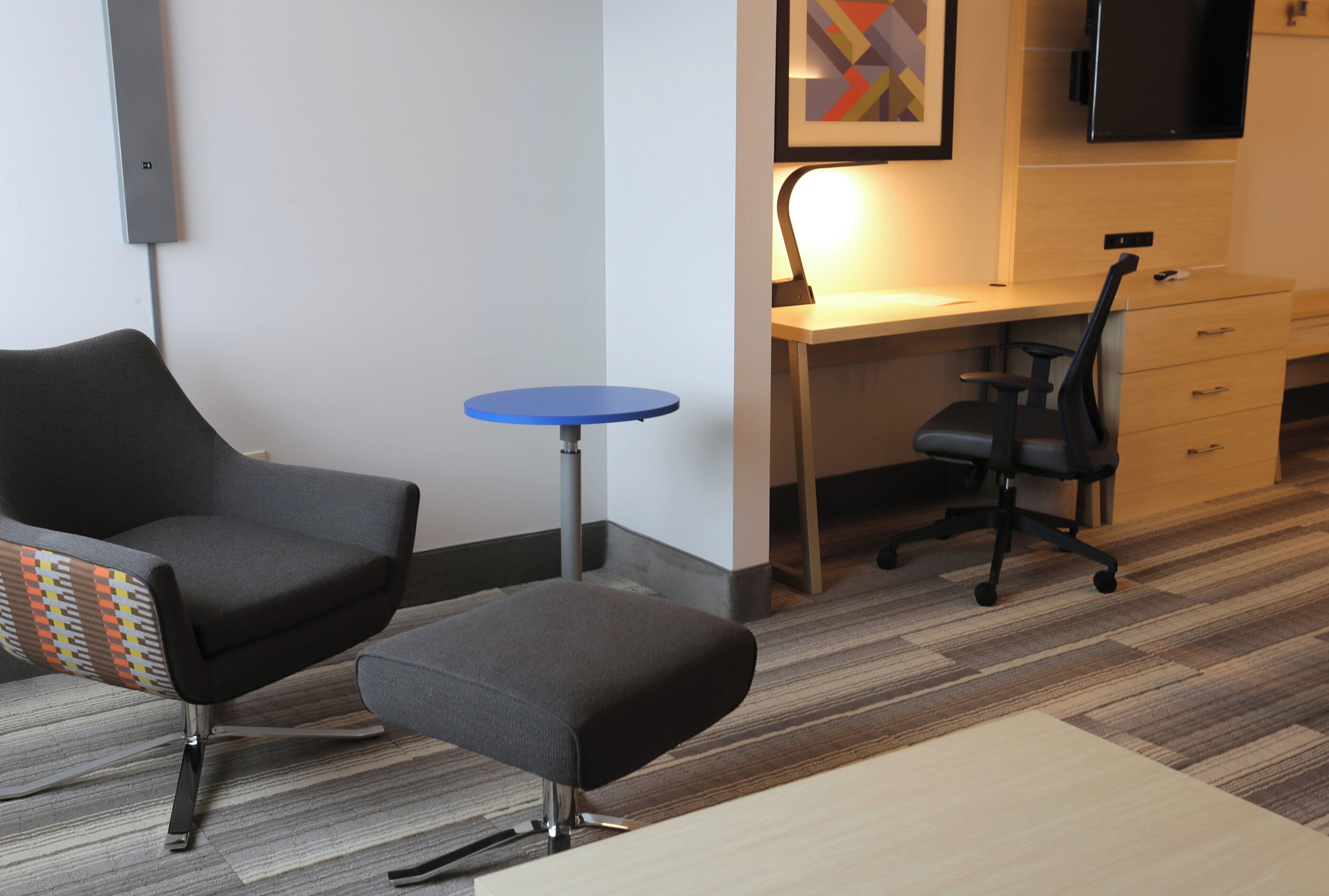 Une chambre d’hôtel moderne avec un fauteuil gris, une petite table bleue, un bureau avec une chaise et un téléviseur à écran plat fixé au mur.