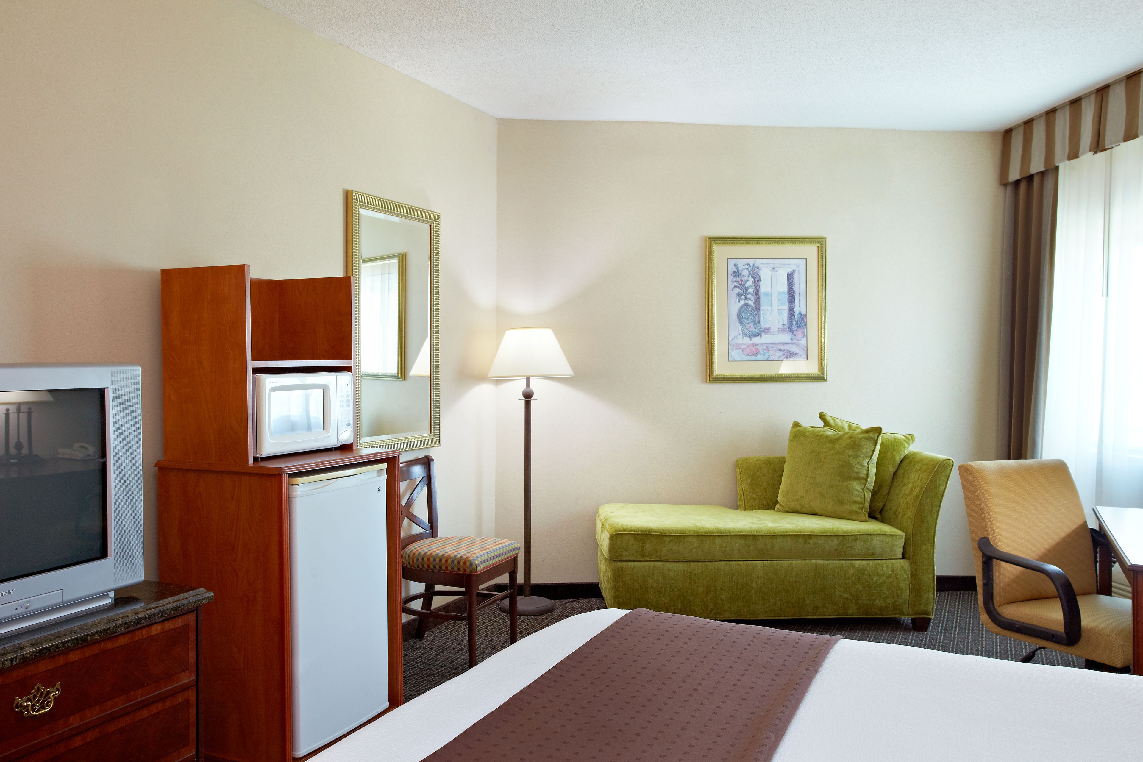 A hotel room with a bed, a desk, a chair, a television, a microwave, a refrigerator, a lamp, and a framed picture on the wall.