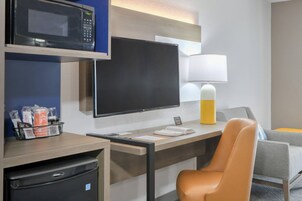 A hotel room with a desk, a microwave, a refrigerator, a TV, a lamp, and a sofa.