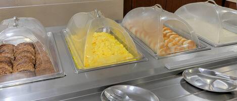 A buffet with covered food containers including what appears to be scrambled eggs, pastries, and possibly a meat dish.