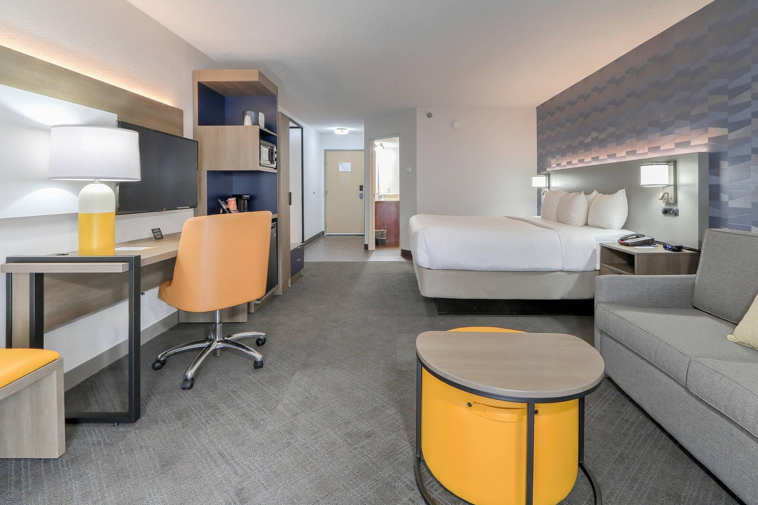 A modern hotel room with a bed, a sofa, a desk with a chair, a small table, and a TV.