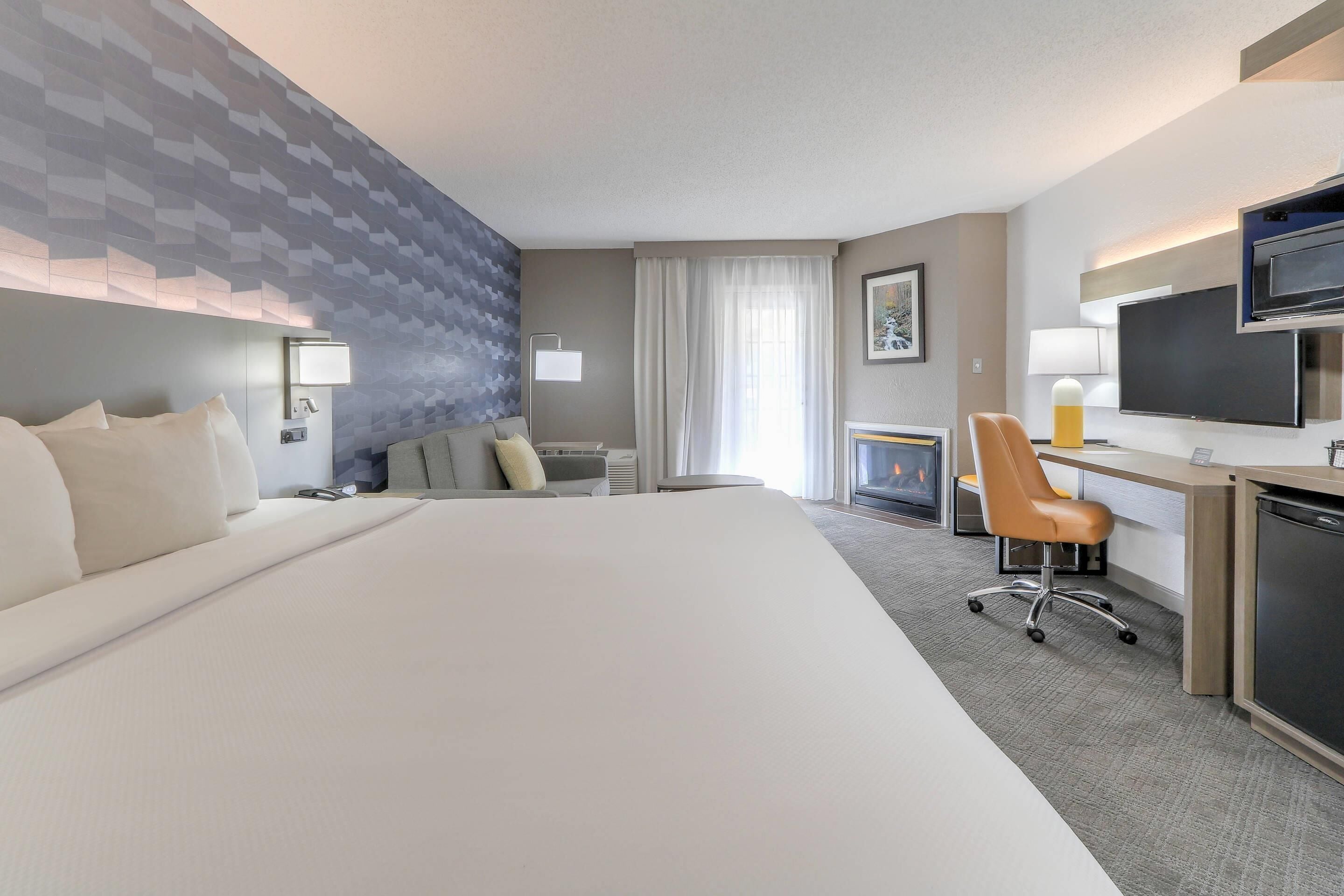 A hotel room with a large bed, a desk with a chair, a television, and a fireplace.