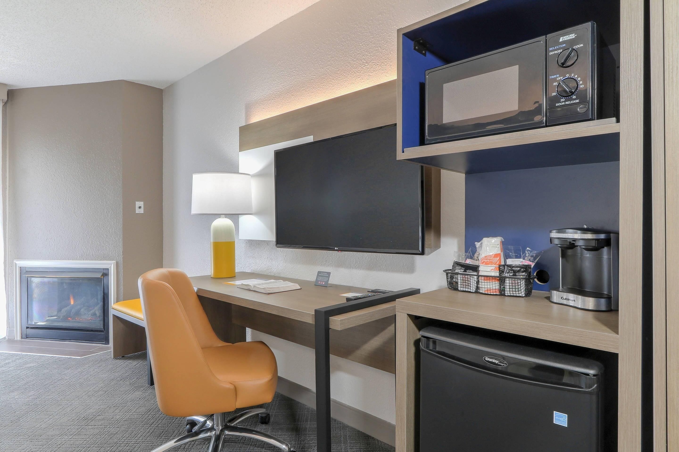 A hotel room with a desk, chair, television, microwave, coffee maker, and a mini-fridge.