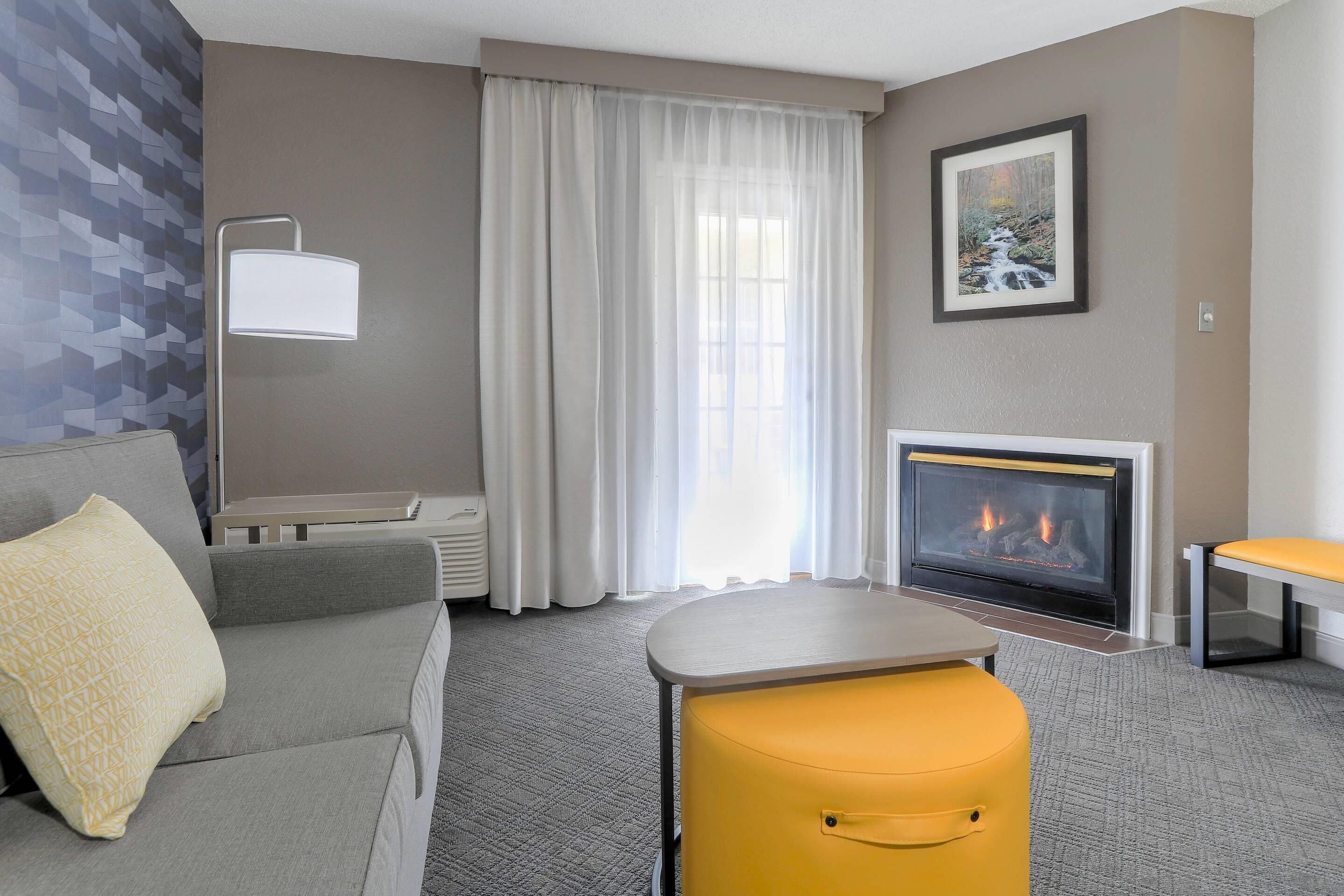 A modern hotel room with a grey sofa, a yellow ottoman, a fireplace, and a large window with sheer curtains.