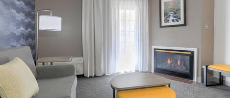 A modern hotel room with a grey sofa, a yellow ottoman, a fireplace, and a large window with sheer curtains.