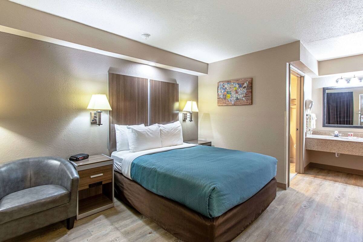 Ein Hotelzimmer mit Bett, Nachttischlampen, einem Sessel, einem Schreibtisch, einem Bild und einem Badezimmerspiegel.
