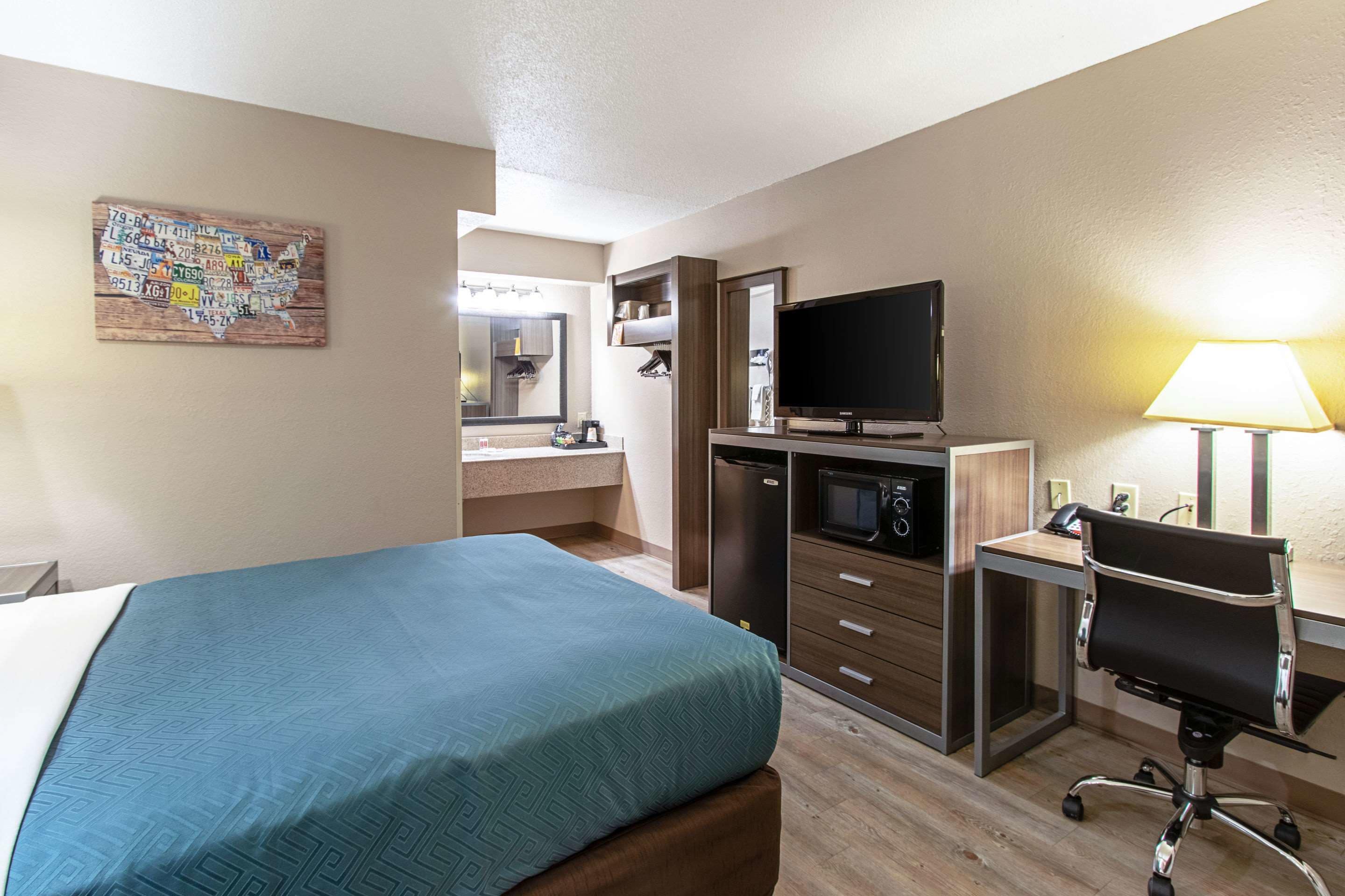 Habitación de hotel con cama, escritorio con computadora, televisor, microondas y baño con espejo y lavamanos.