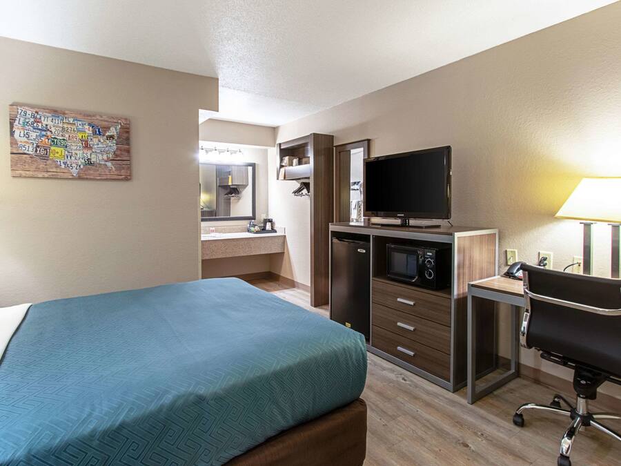 Quarto de hotel com cama, escrivaninha com computador, televisão, micro-ondas e banheiro com espelho e pia.