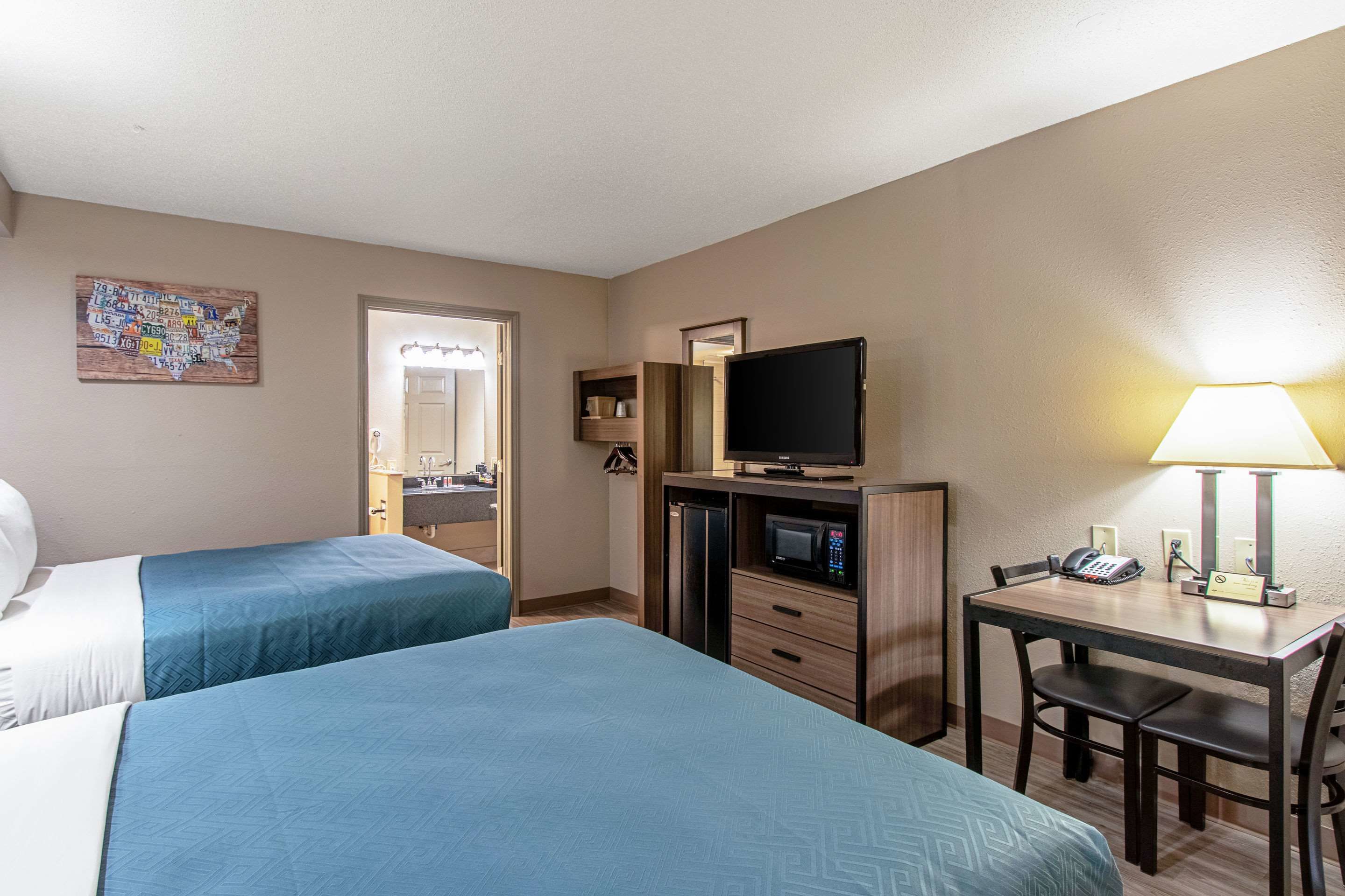 Quarto de hotel com duas camas, uma escrivaninha com luminária, televisão e uma pequena cozinha.