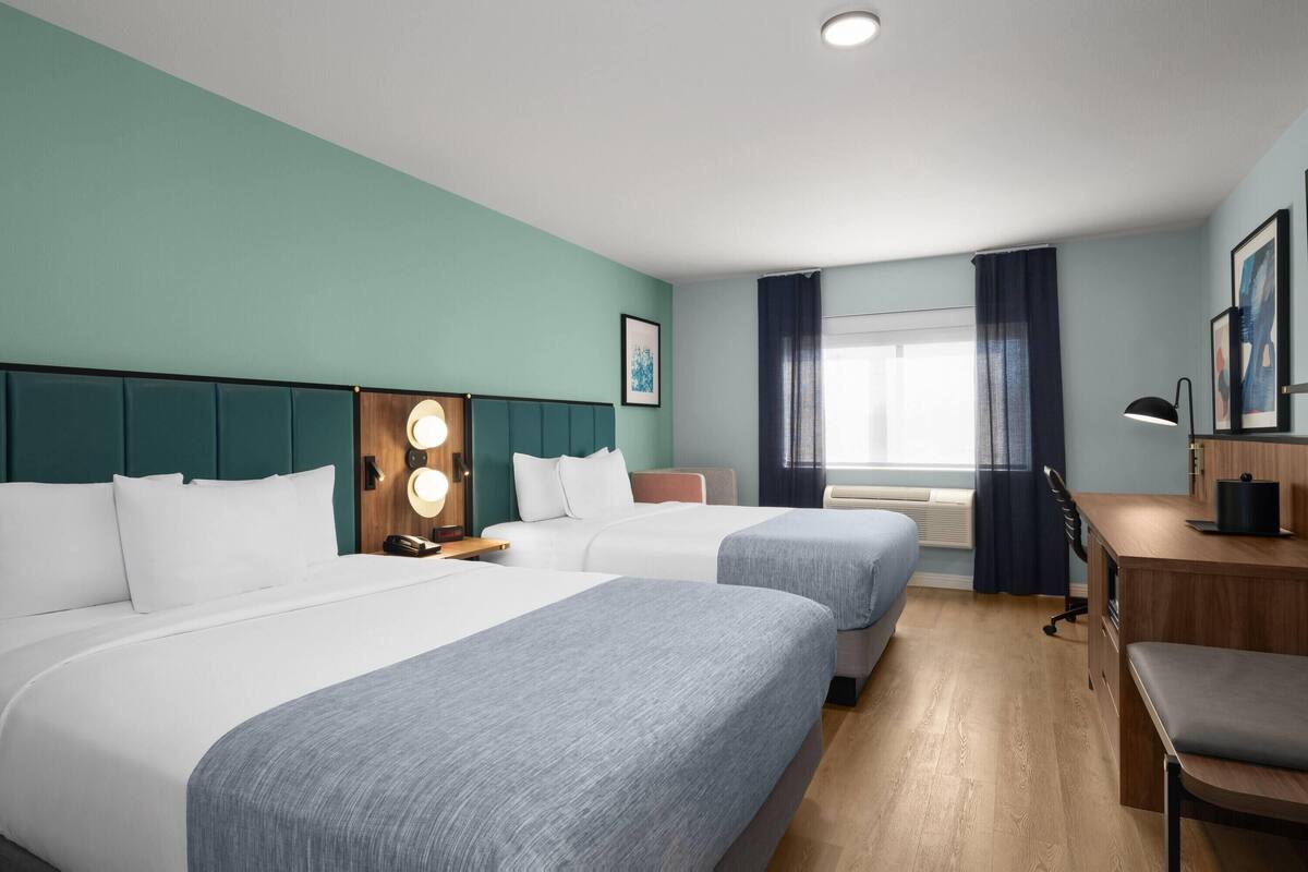 Quarto de hotel com duas camas, uma escrivaninha, uma cadeira, luminária de mesa, janela com cortinas e telefone fixado na parede.