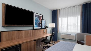Habitación de hotel con cama, escritorio, silla, televisor, aire acondicionado y una obra de arte en la pared.