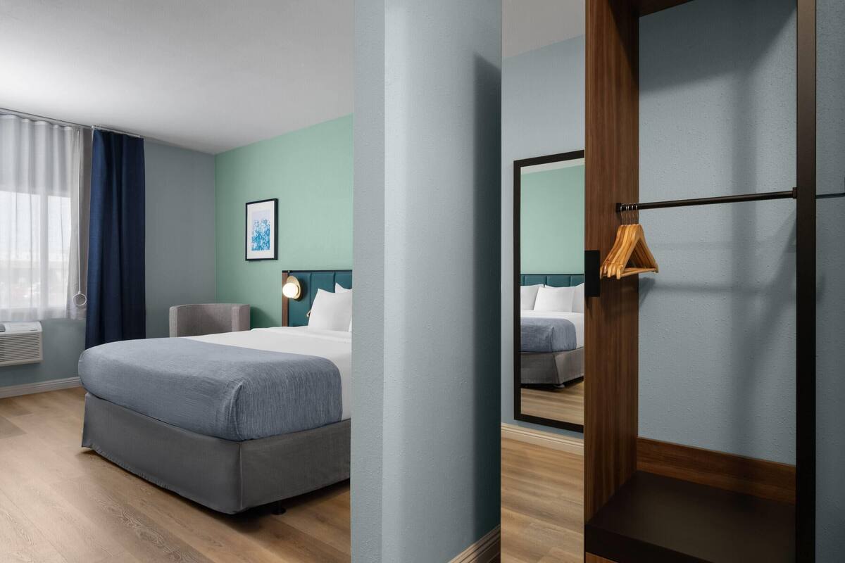 Quarto de hotel com cama, uma cadeira, escrivaninha, espelho e janela com cortinas.