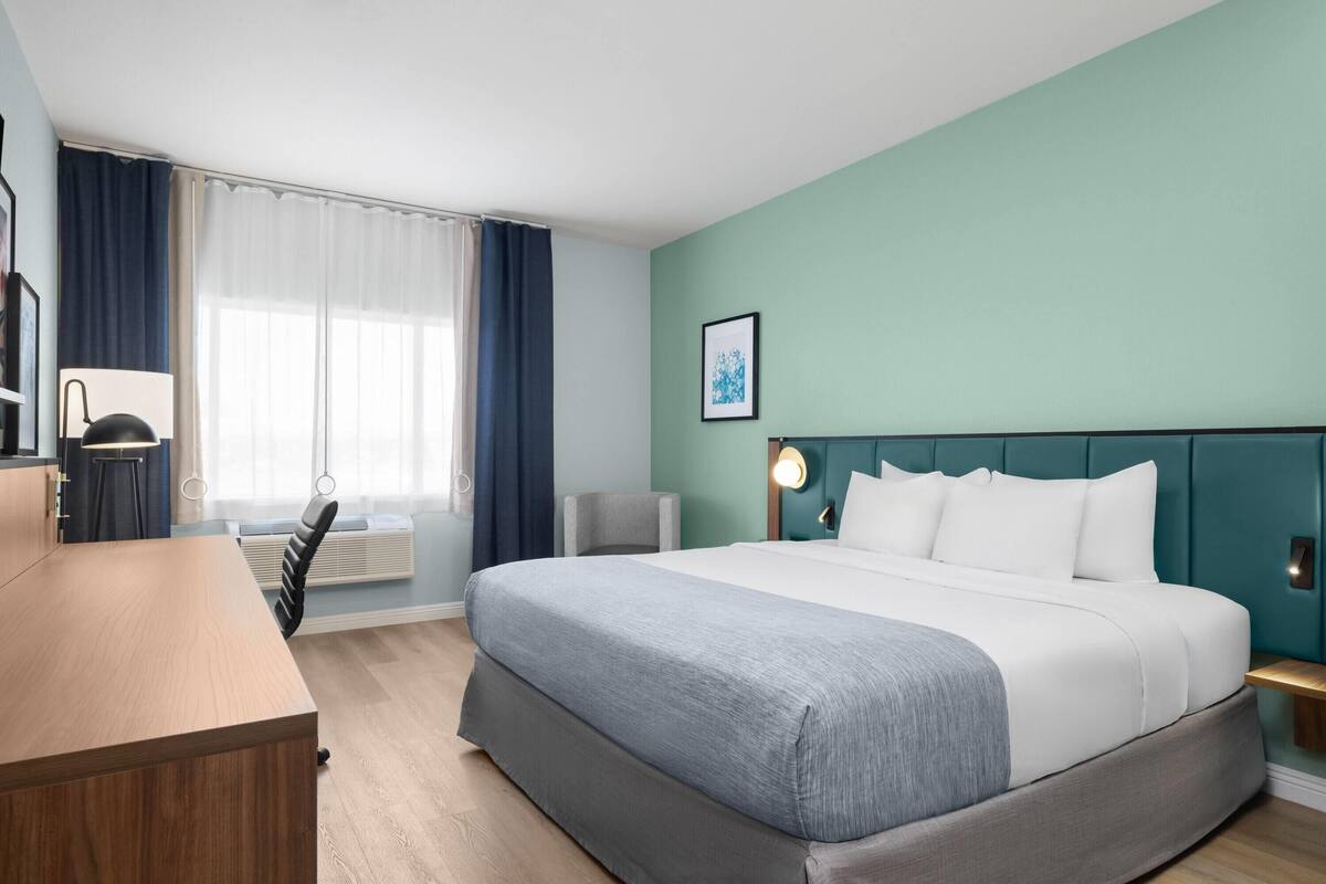 Quarto de hotel com cama, escrivaninha, cadeira, janela com cortinas e luminária na parede.